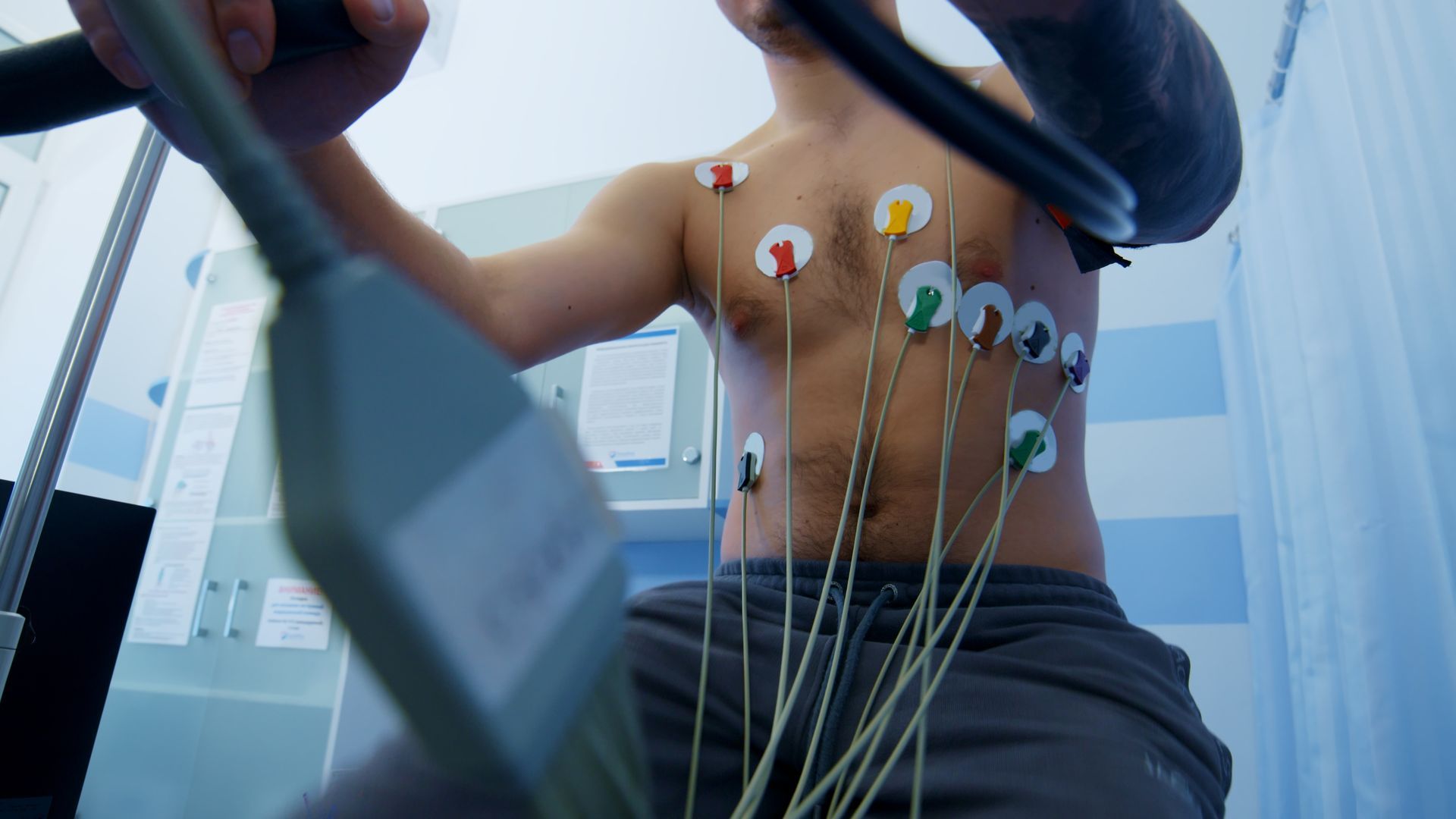 Man with electrodes on his chest, exercising on a stationary bike in a medical setting.