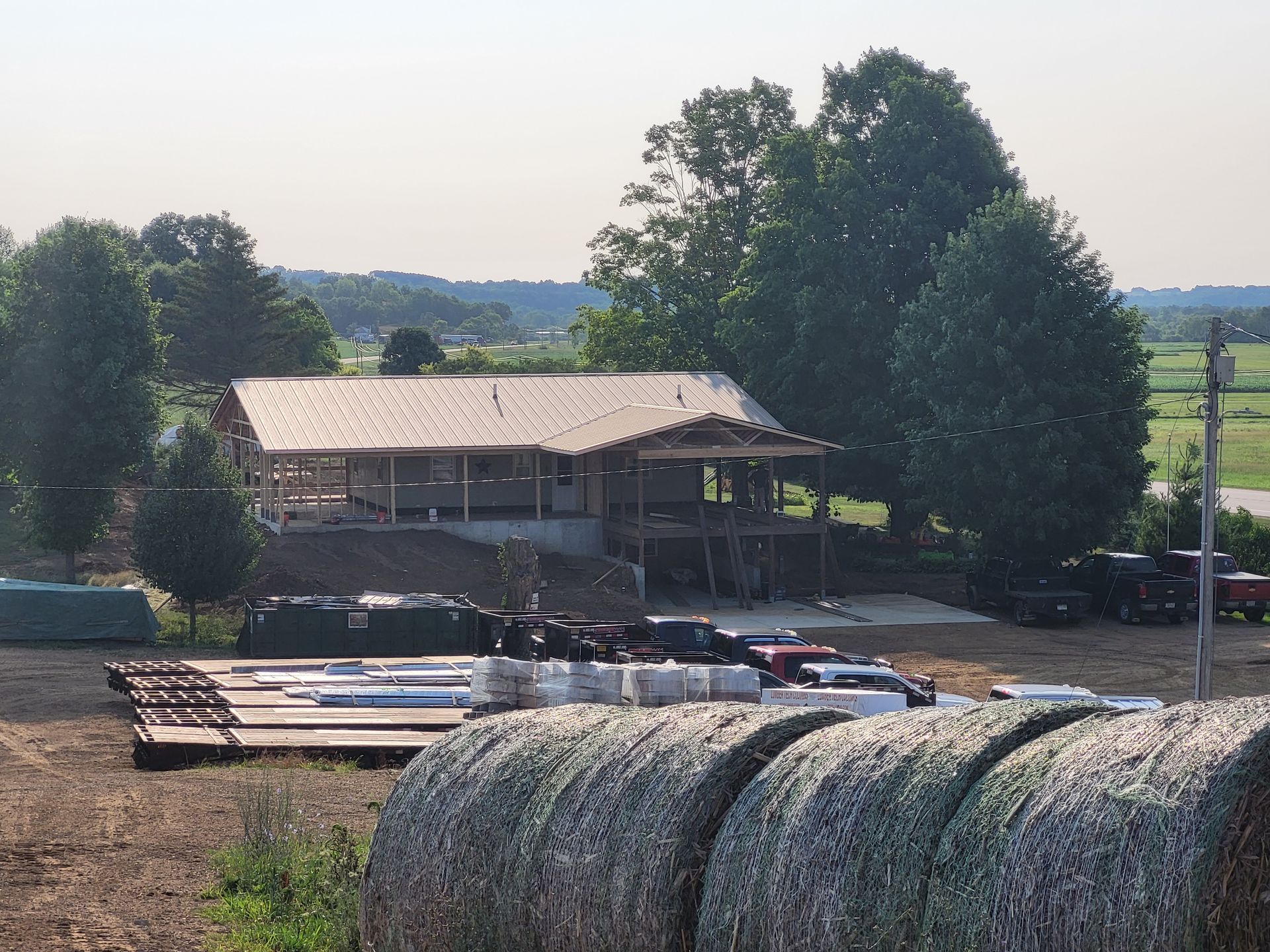 Construction site with a partially built house, hay bales in the foreground, and trees in the background.