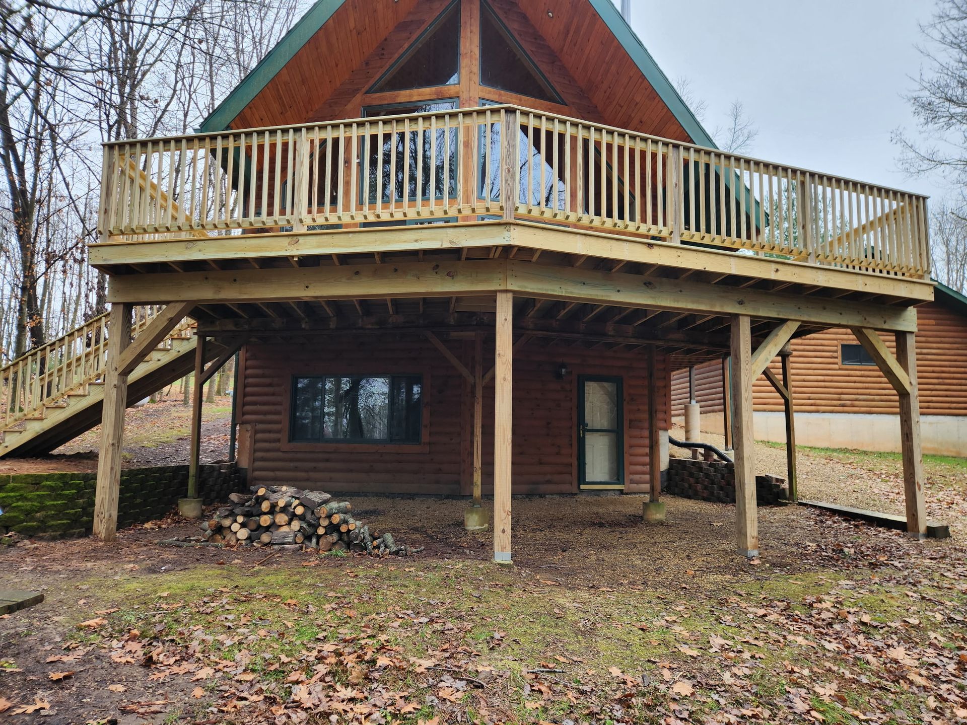 A-frame cabin with a large wooden deck. Log siding, steps to the deck, and a woodpile beneath.
