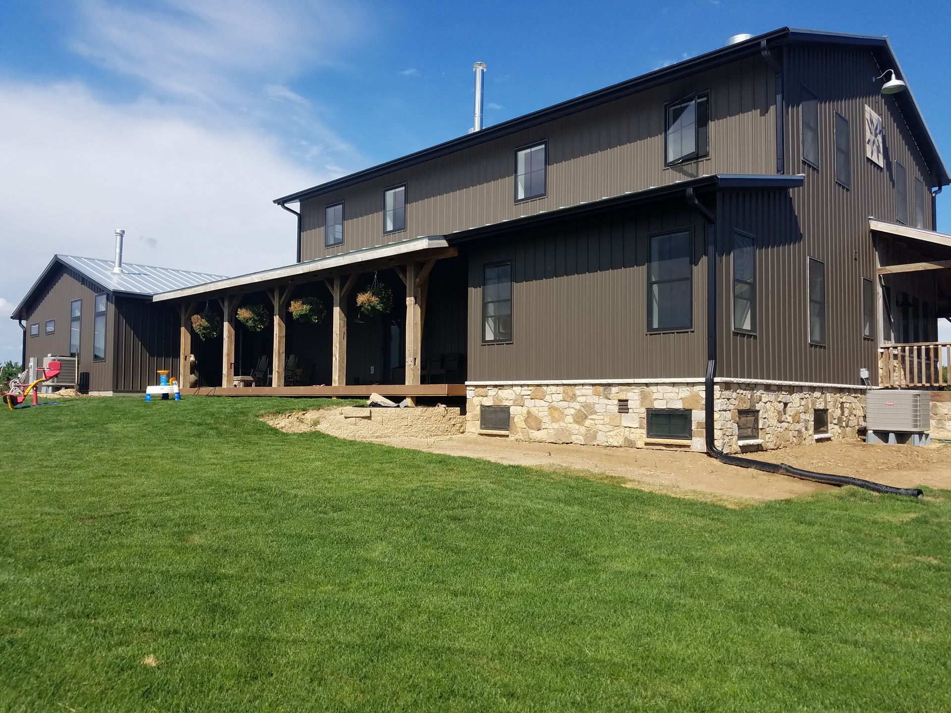 Dark brown barn-style building with a porch, stone foundation, and green lawn on a sunny day.