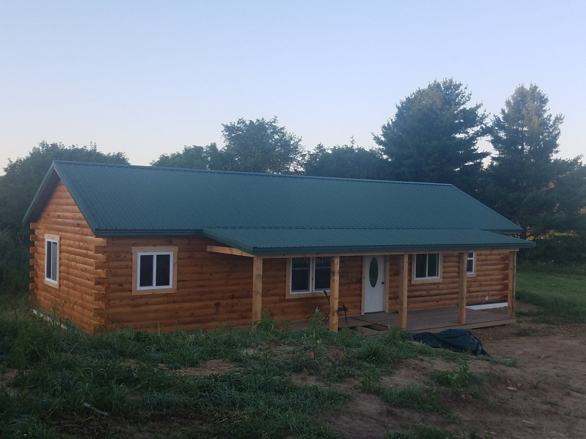 Log cabin with a green roof and porch, set in a grassy field.