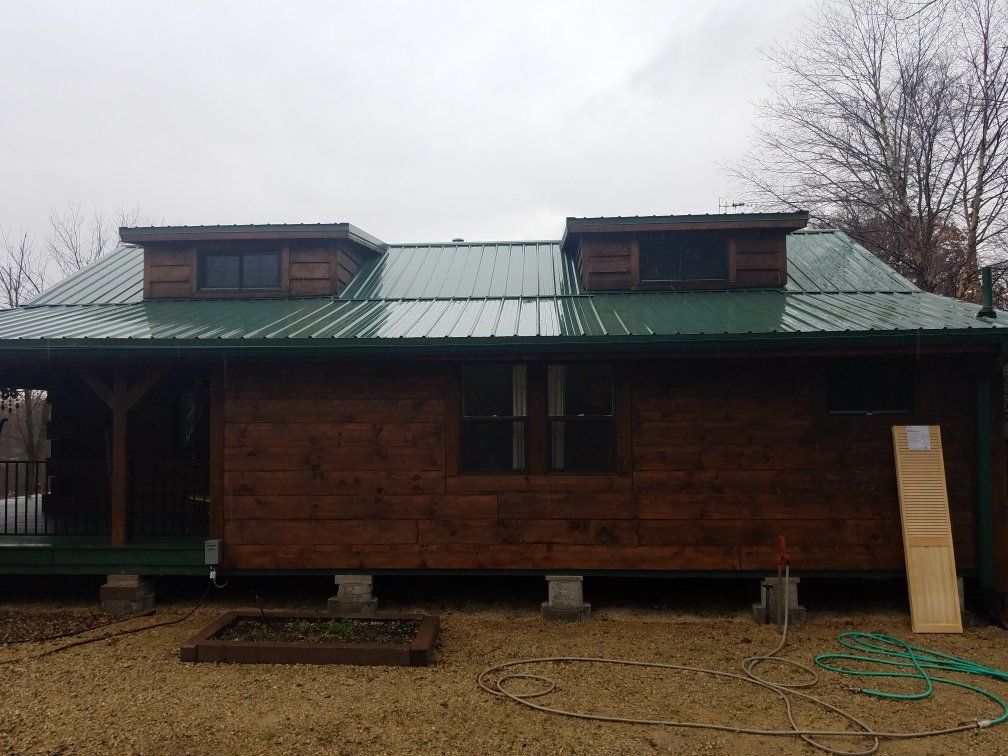 Cabin with green metal roof, dark wood siding, two dormers, and porch. Cloudy day.