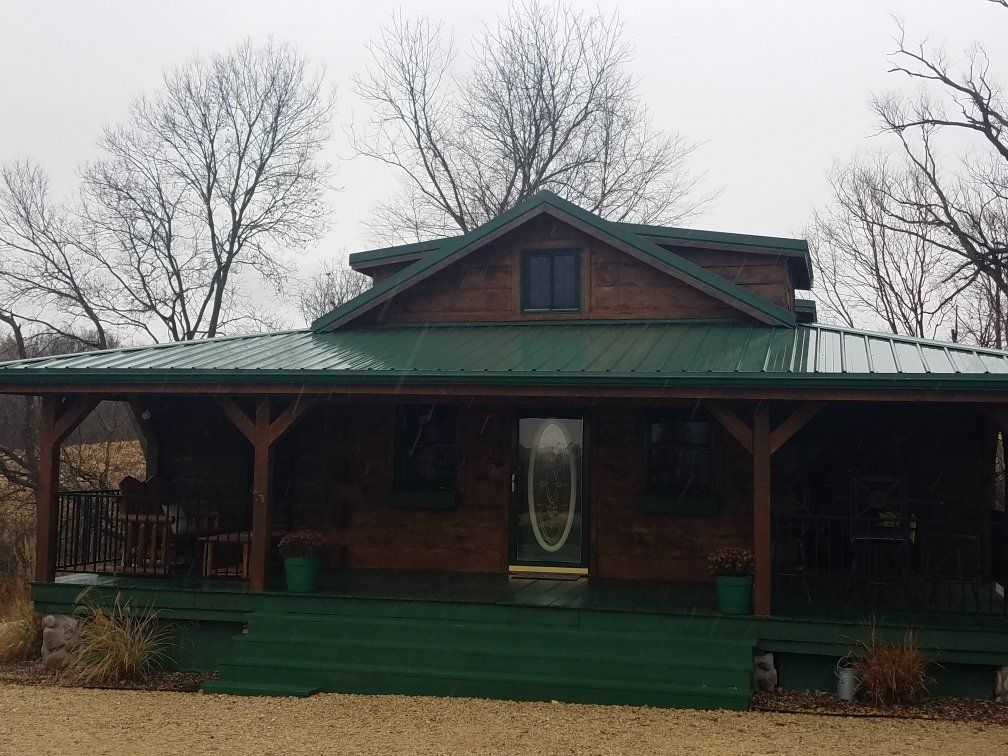 Brown cabin with green roof and porch, on a cloudy day. Bare trees in the background.