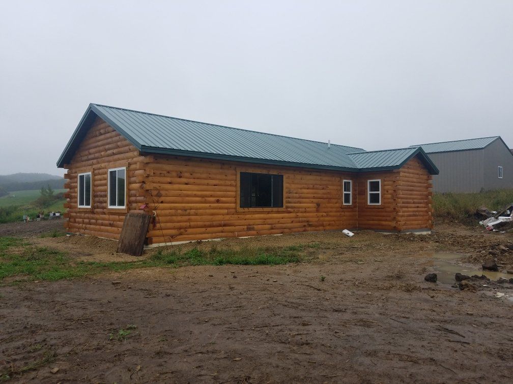 Log cabin with teal roof and white-framed windows, set on muddy ground under a cloudy sky.