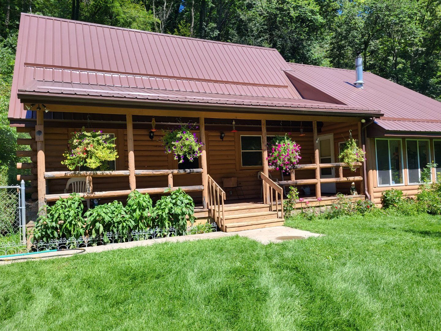 Log cabin with brown roof, porch, and hanging flowers, set in a grassy yard.