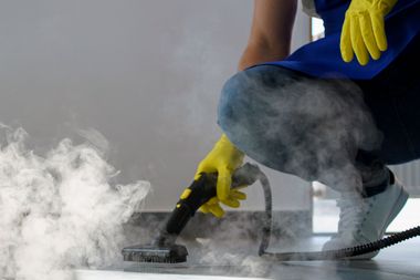 Worker in yellow gloves steam-cleans a floor, with white steam rising around the nozzle.