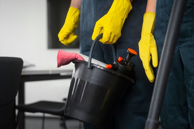 Person in yellow gloves carrying a black bucket with cleaning supplies near a chair and table