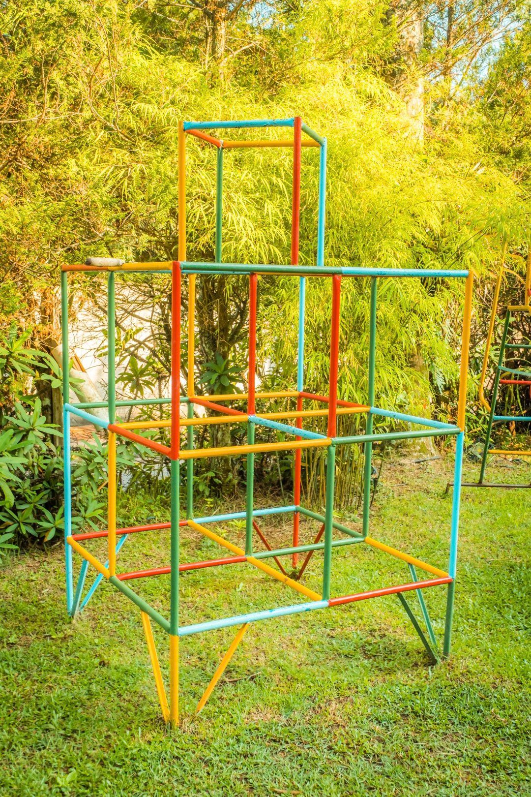 Estrutura metálica colorida de um parque infantil em um gramado, com barras amarelas, vermelhas, azuis e verdes em meio às árvores.