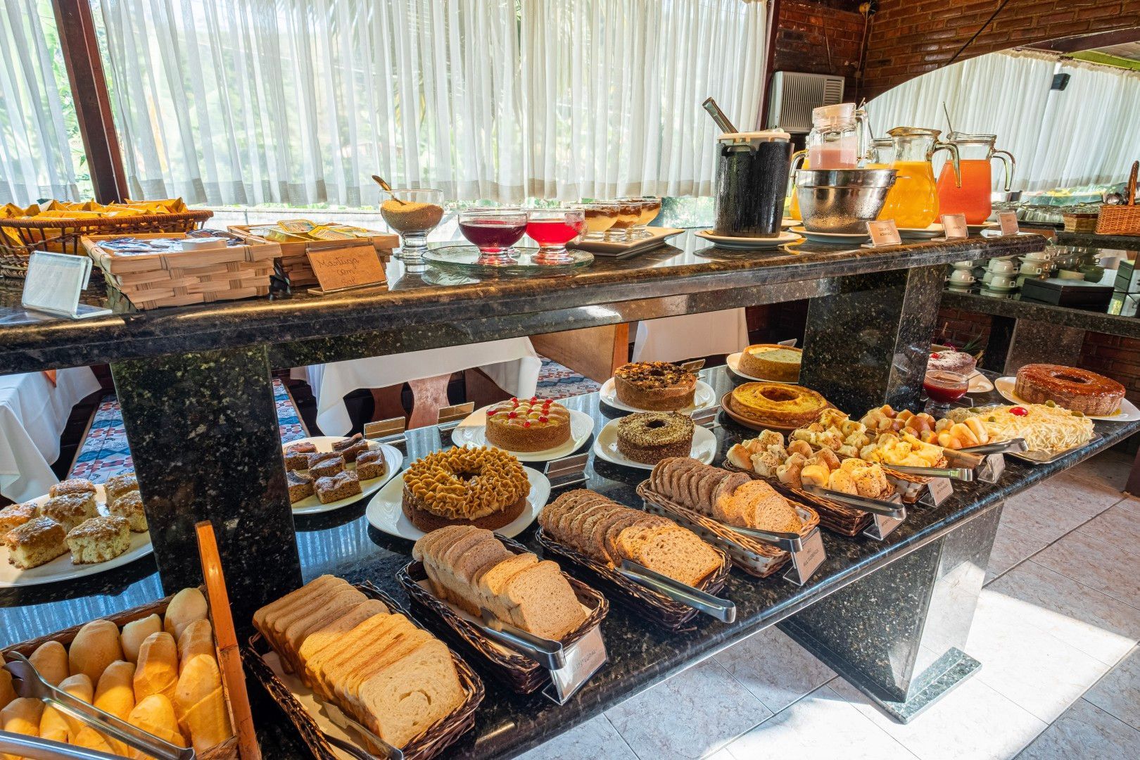 Mesa de buffet com uma variedade de doces, pães, frutas e bebidas em uma área de jantar iluminada.