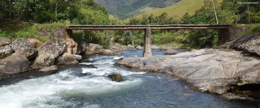 Pequena ponte sobre um rio rochoso em uma paisagem montanhosa verdejante.