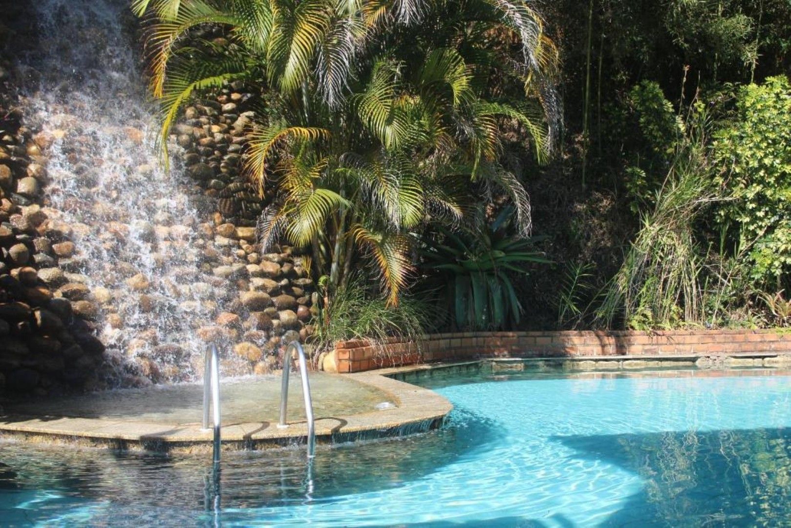 Piscina de resort com cascata, palmeiras e água azul em um ambiente tropical.