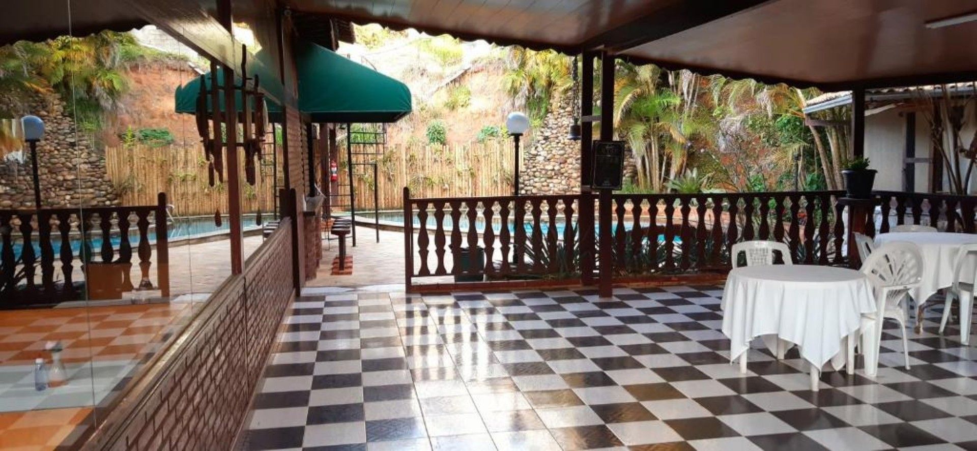Pátio ao ar livre com piso xadrez preto e branco, mesas e cadeiras brancas e vista para a mata.