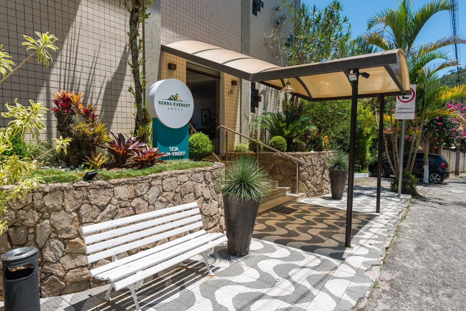 Entrada de café ao ar livre com parede de pedra, banco branco, vasos de plantas e toldo ao longo de um caminho de paralelepípedos.