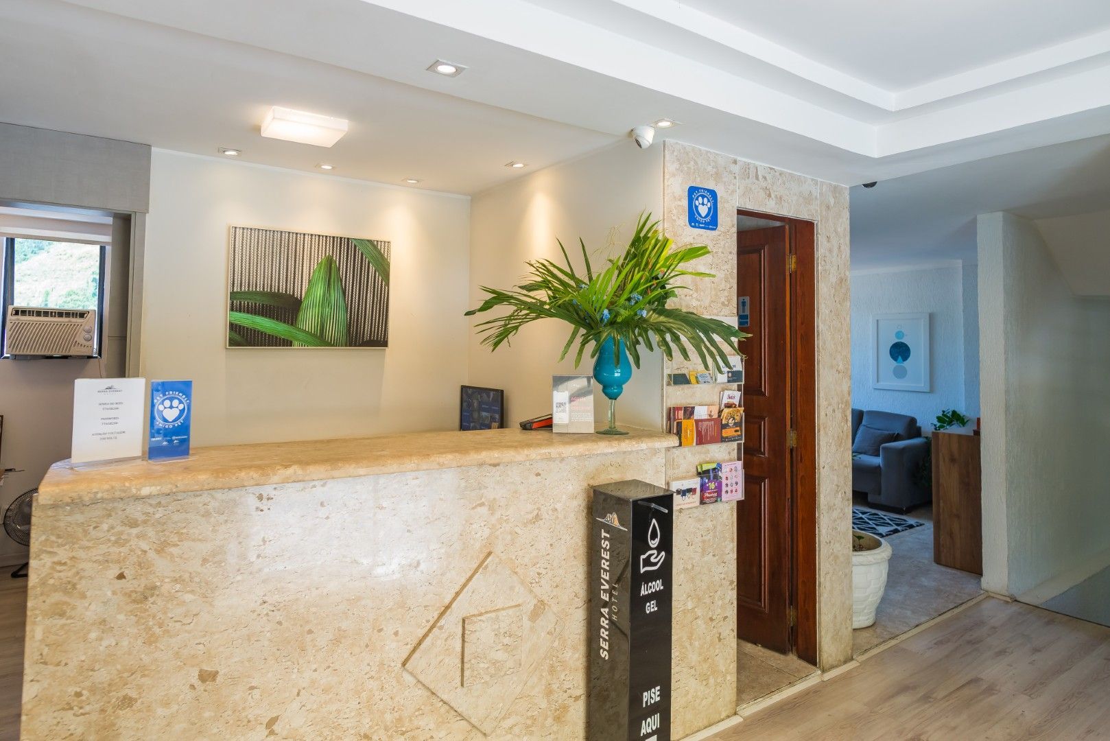 Balcão de recepção do hotel em um saguão iluminado com obras de arte, plantas e um corredor ao fundo.