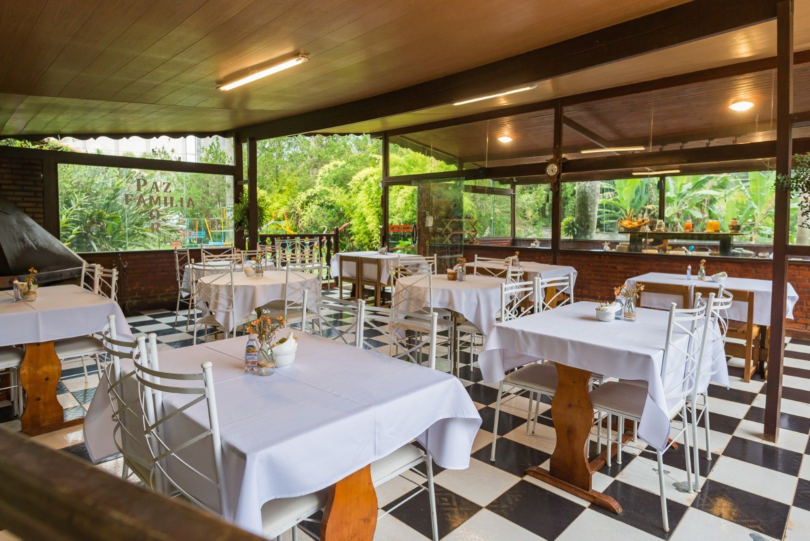 Restaurante ao ar livre com mesas de toalha branca e piso xadrez preto e branco com vista para a vegetação.