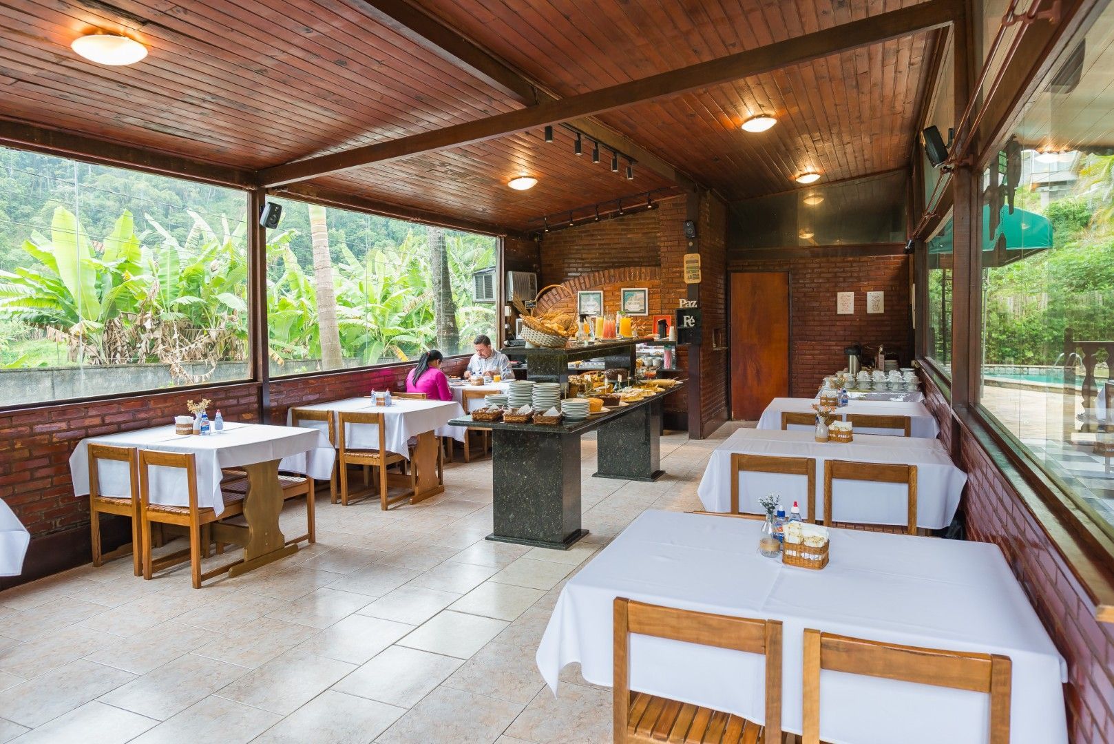 Restaurante ao ar livre com mesas de madeira, toalhas de mesa brancas e iluminação aconchegante em um ambiente tropical.