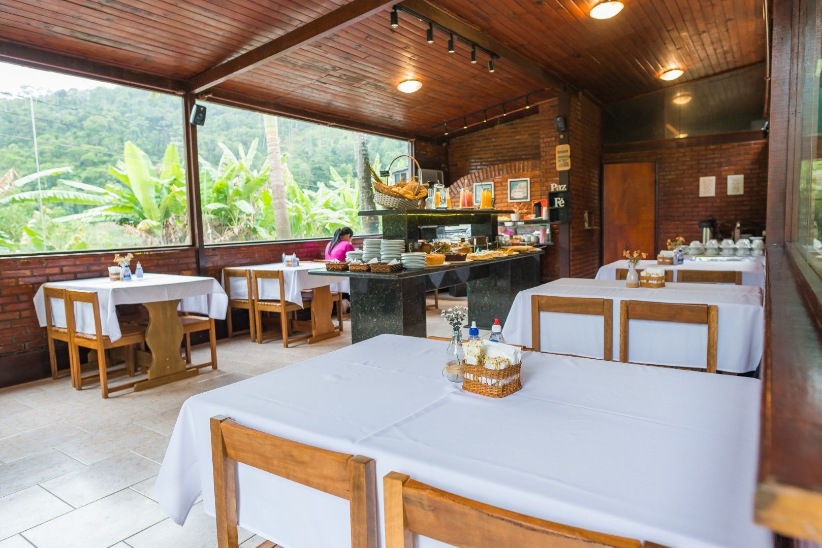 Restaurante ao ar livre com mesas de madeira, toalhas de mesa brancas e balcão de buffet com vista para colinas verdejantes.