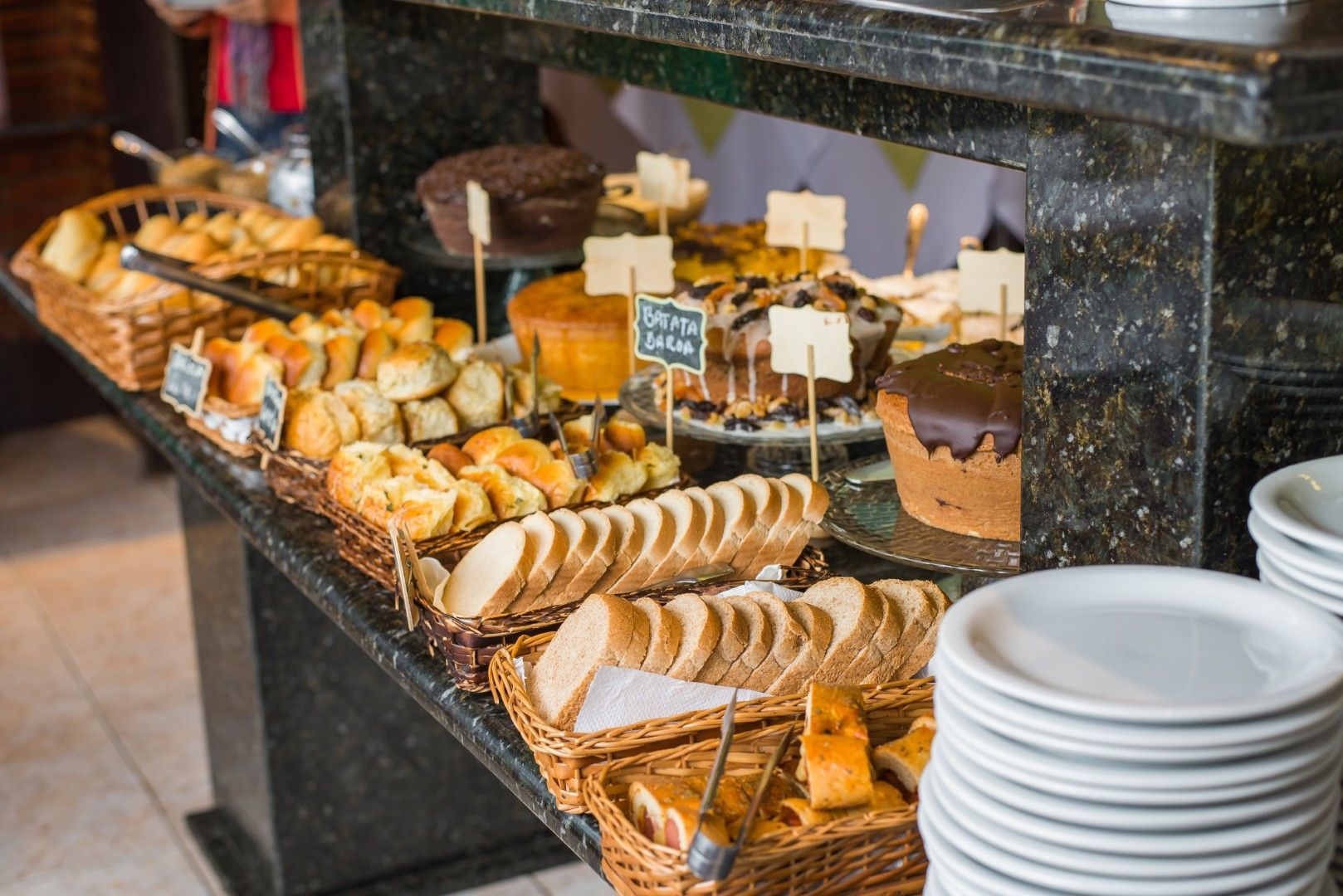 Buffet de sobremesas com uma variedade de doces e bolos em um balcão escuro ao lado de pratos brancos empilhados.