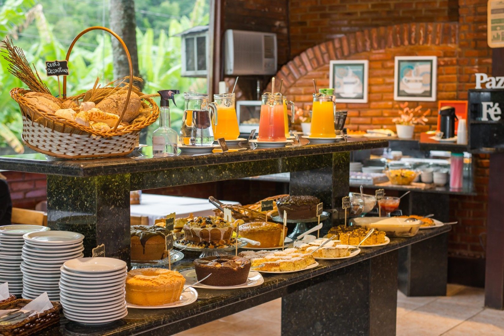 Buffet de hotel com doces, frutas e sucos em uma área de refeições com paredes de tijolos.