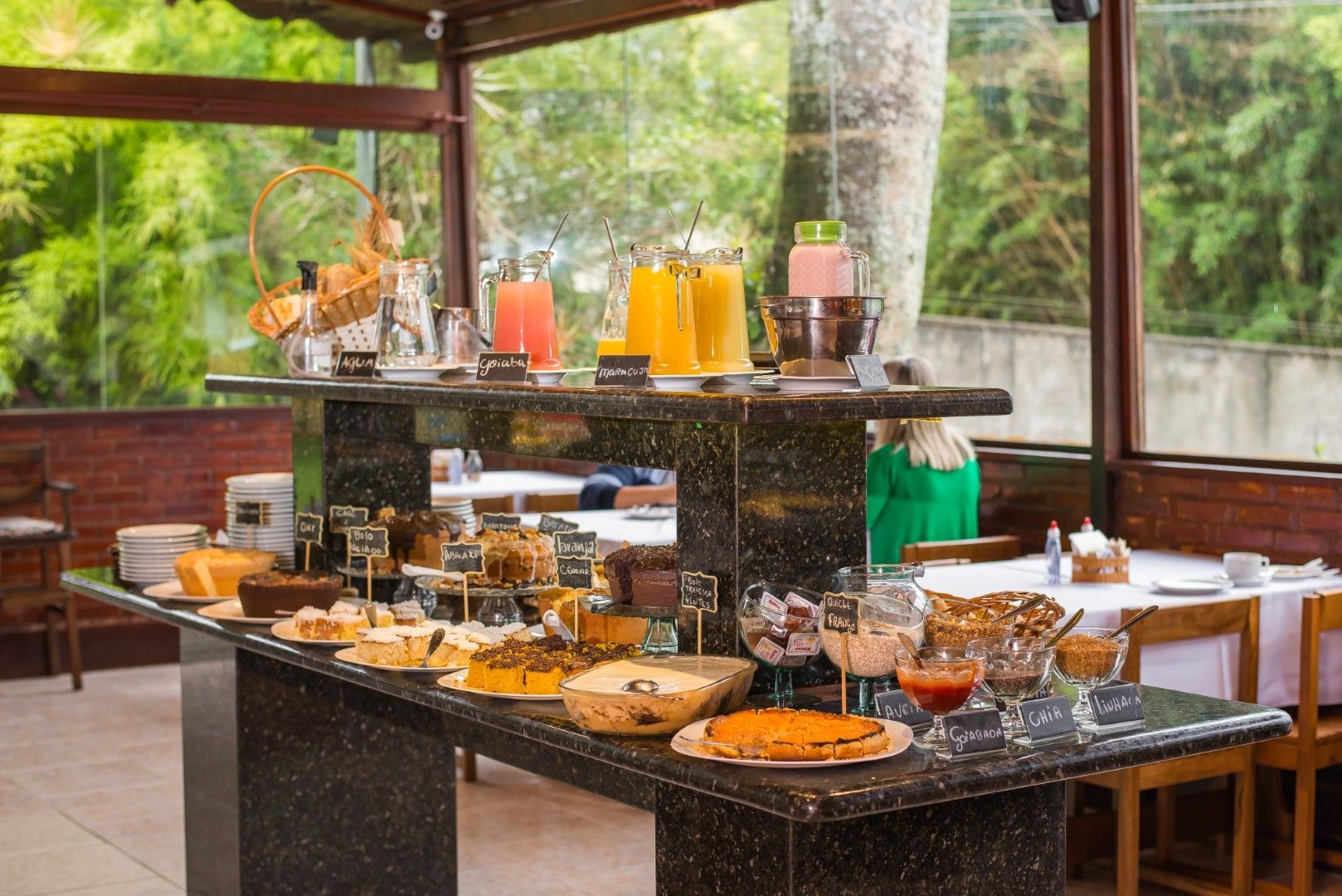 Mesa de buffet com bebidas, doces e frutas em um ambiente luminoso com áreas internas e externas para refeições.