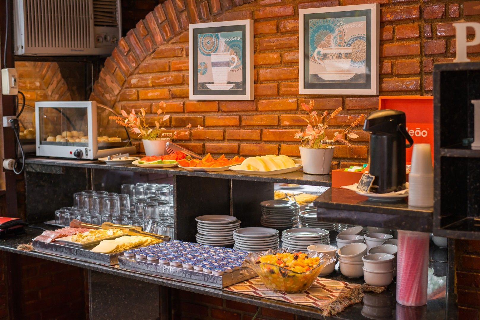 Mesa de buffet com doces, tigelas, cafeteira e copos em um restaurante com paredes de tijolos.