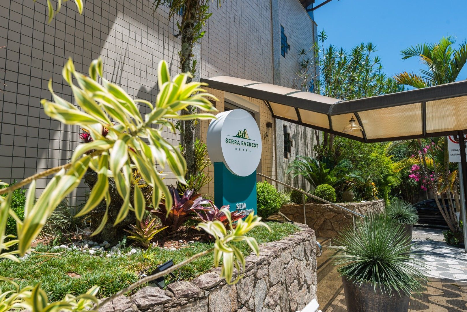 Entrada de jardim ensolarada com placa, muro de pedra e plantas tropicais ao lado de um edifício.