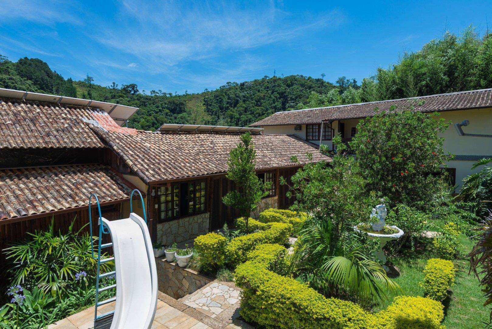 Pátio com edifícios de telhado de telha, jardim verdejante e um escorregador branco sob um céu azul brilhante.