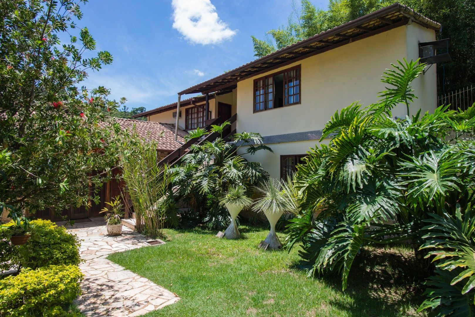 Casa ensolarada com caminho de jardim, gramado verde e plantas tropicais.