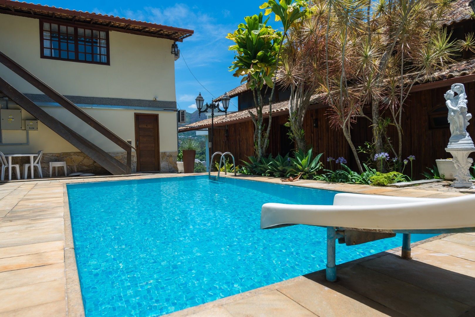 Piscina ensolarada no quintal com água azul, trampolim, pátio e fachada da casa.