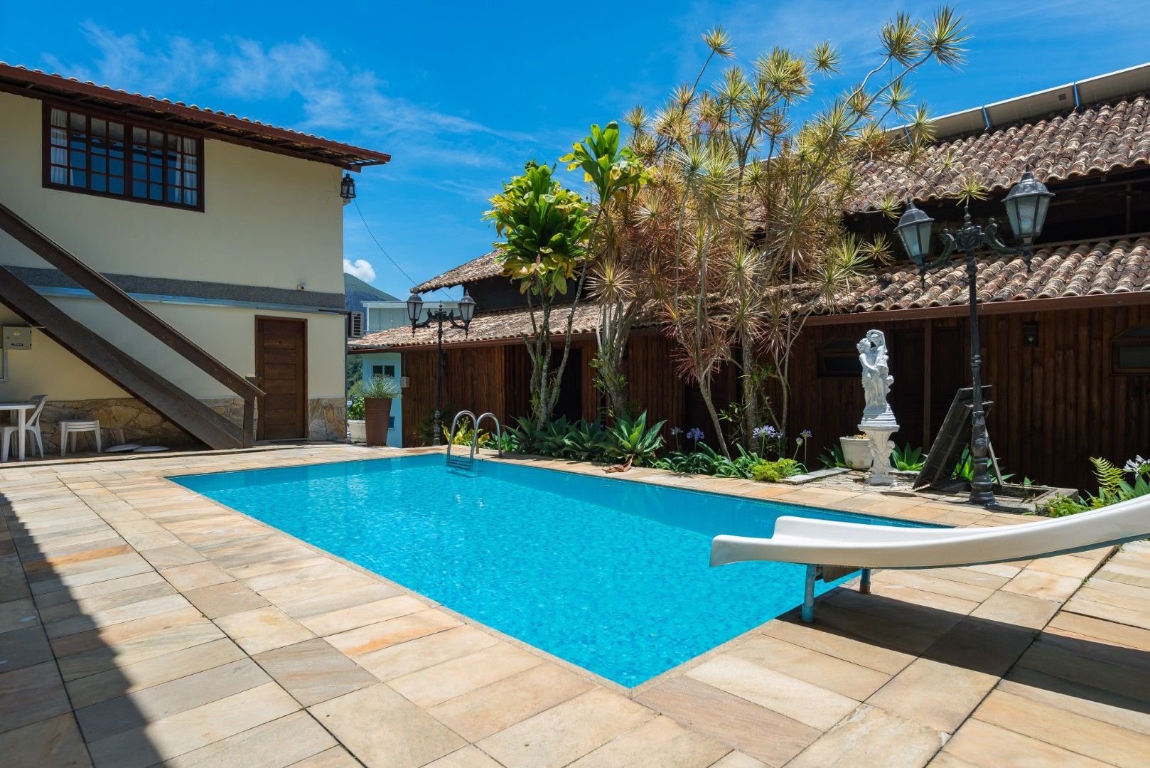 Pátio ensolarado com piscina azul, piso de cerâmica, espreguiçadeiras e construções em estilo espanhol sob um céu claro.