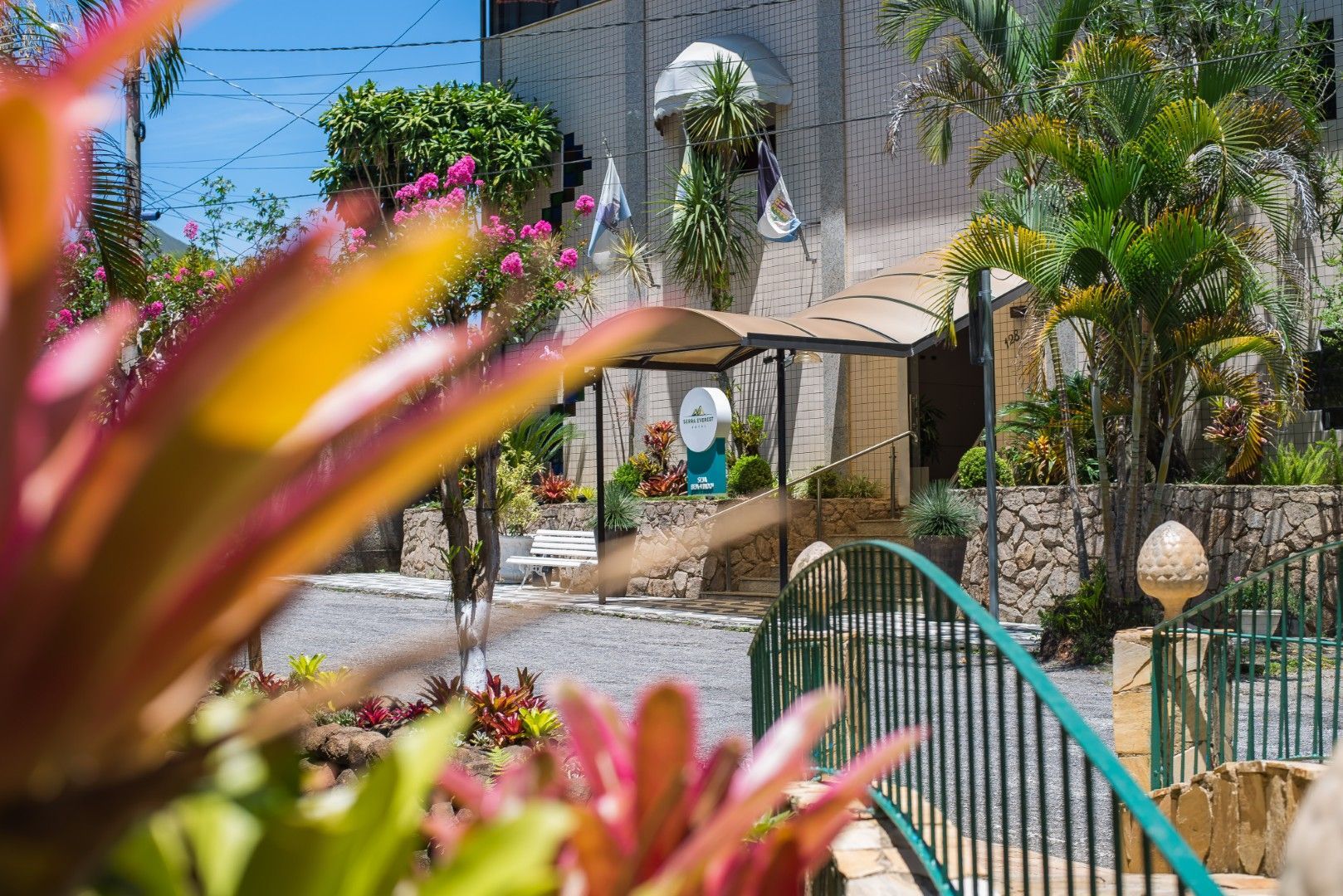 Pátio de jardim tropical com palmeiras, flores cor-de-rosa, um portão azul e um edifício de pedra branca banhado de sol.