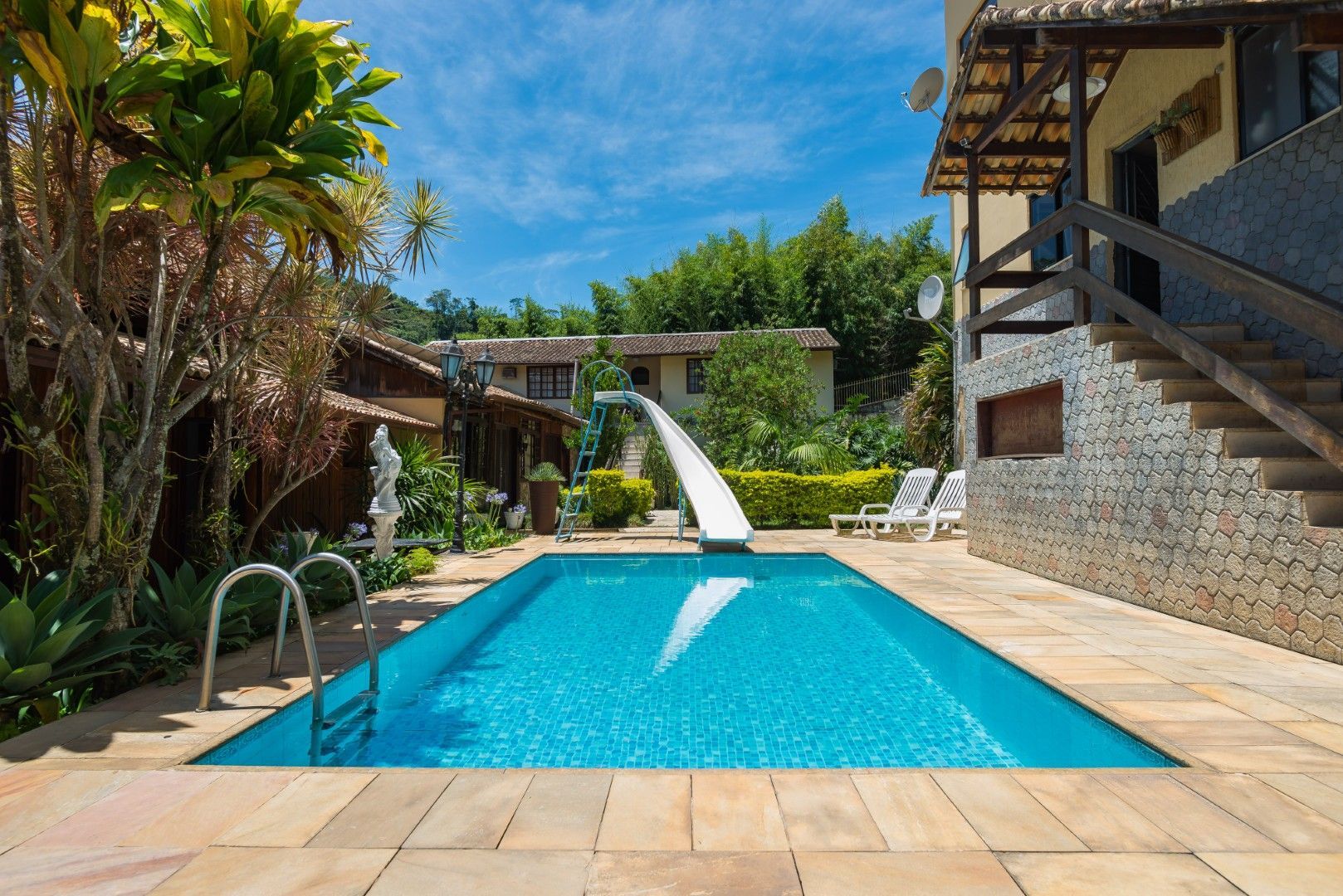 Piscina de resort com água azul, palmeiras e um toboágua ao lado de construções de pedra sob um céu ensolarado.