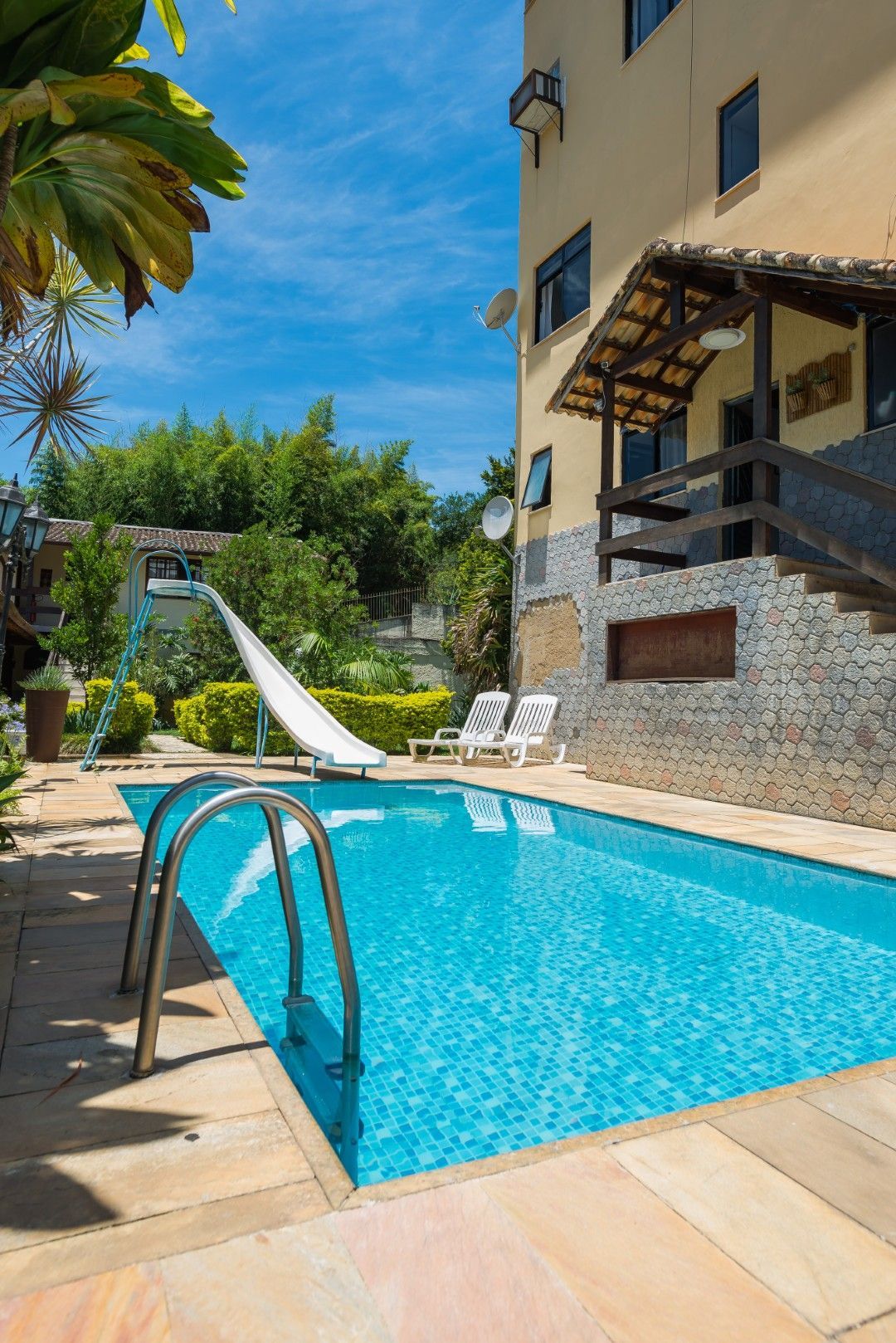 Piscina no quintal ao lado de um prédio bege, com escorregador, escada e palmeiras sob um céu azul ensolarado.