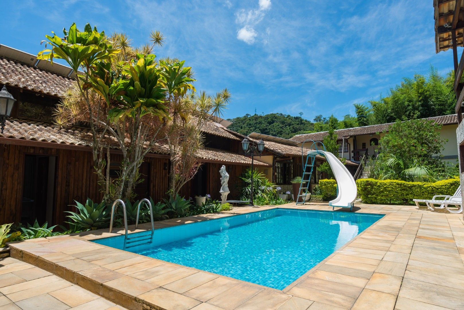 Pátio do resort com piscina azul, terraço de pedra, plantas tropicais e um toboágua branco sob um céu ensolarado.