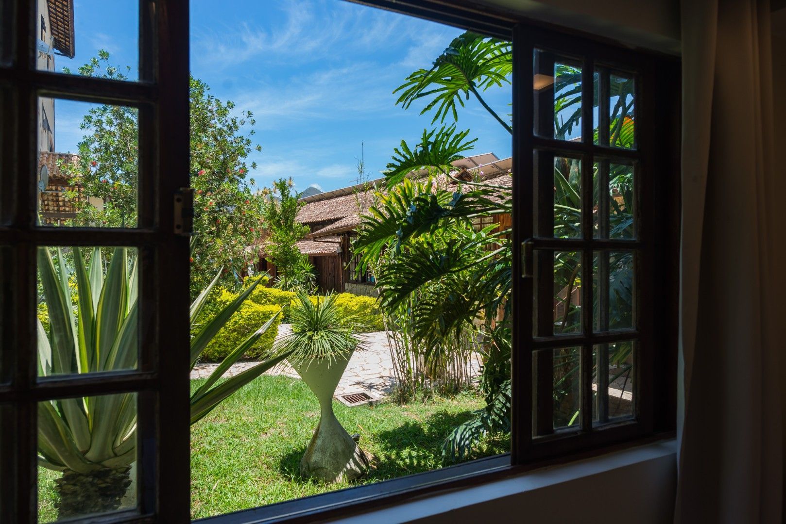 Vista através de uma janela aberta para um jardim ensolarado com palmeiras, grama e uma casa de telhado de palha ao longe.