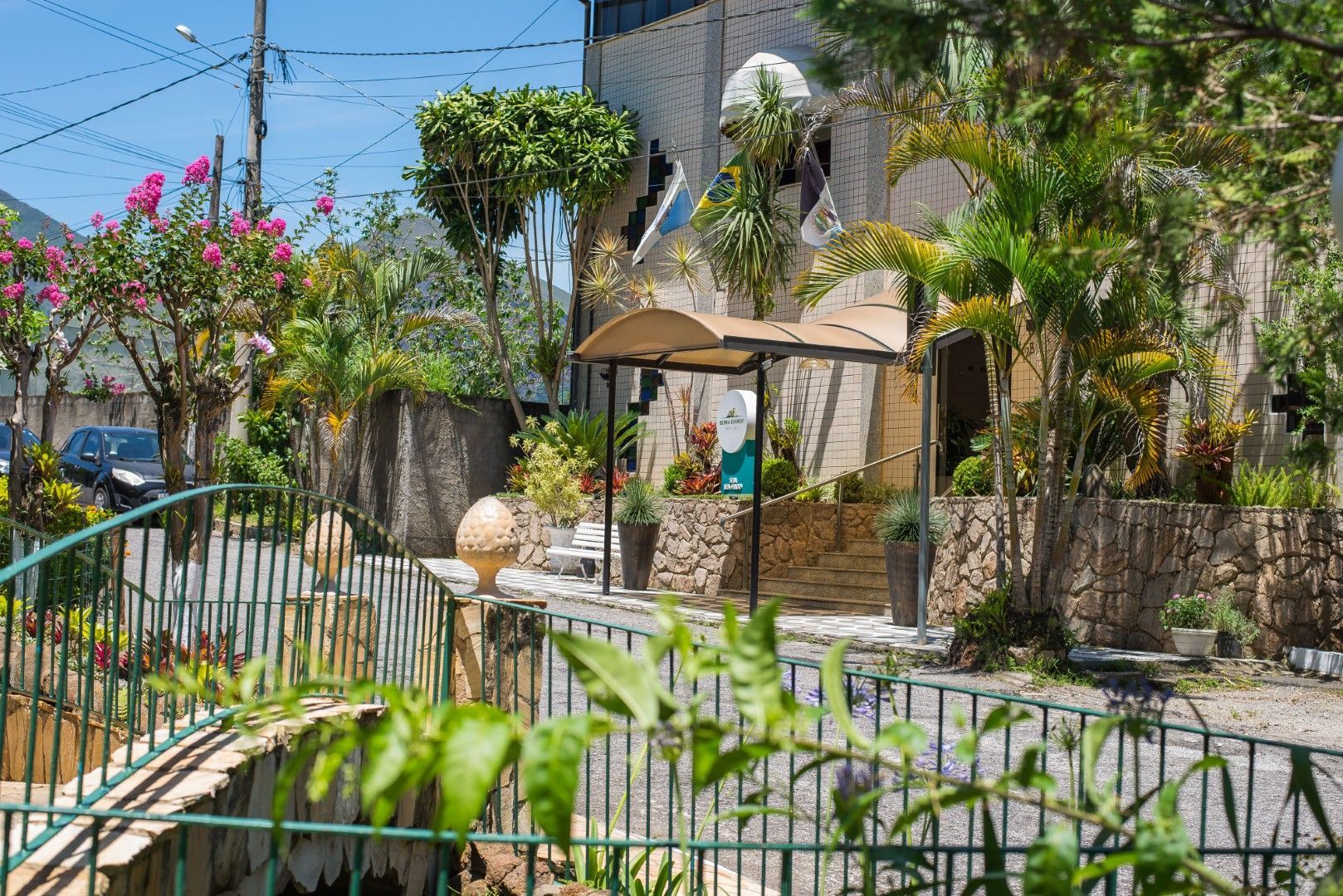 Pátio ensolarado com palmeiras, muros de pedra, guarda-sóis brancos e grades verdes junto à piscina.