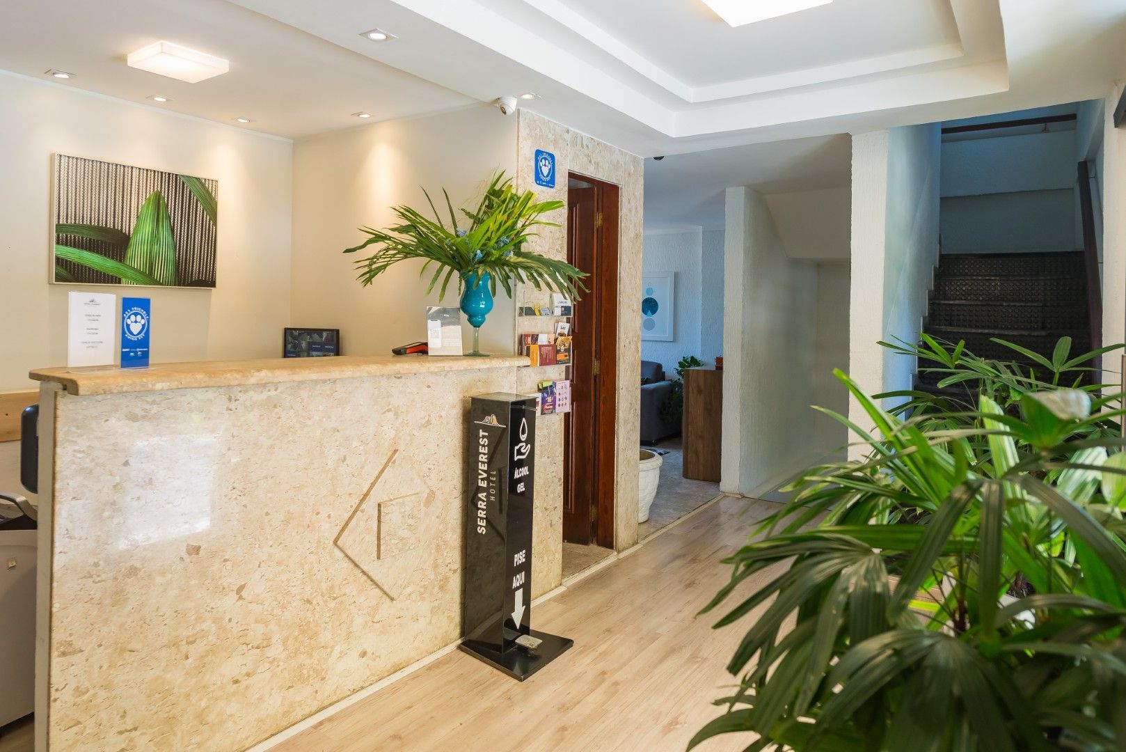 Hall de entrada moderno de hotel com balcão de recepção em mármore, plantas em vasos e escadas ao fundo.