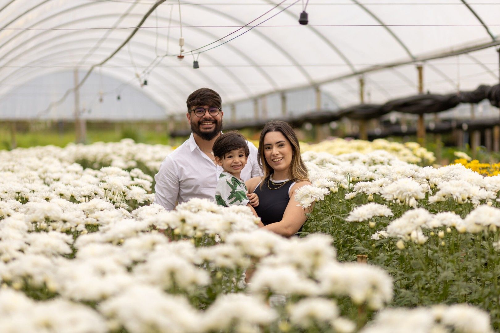 Três pessoas sorrindo em uma estufa de flores, rodeadas por flores brancas.
