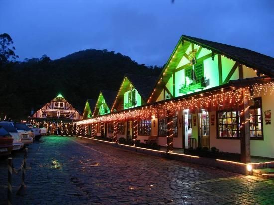 Lojas iluminadas em estilo chalé alinham-se ao entardecer numa rua de paralelepípedos molhada, com telhados iluminados de verde e uma montanha ao fundo.