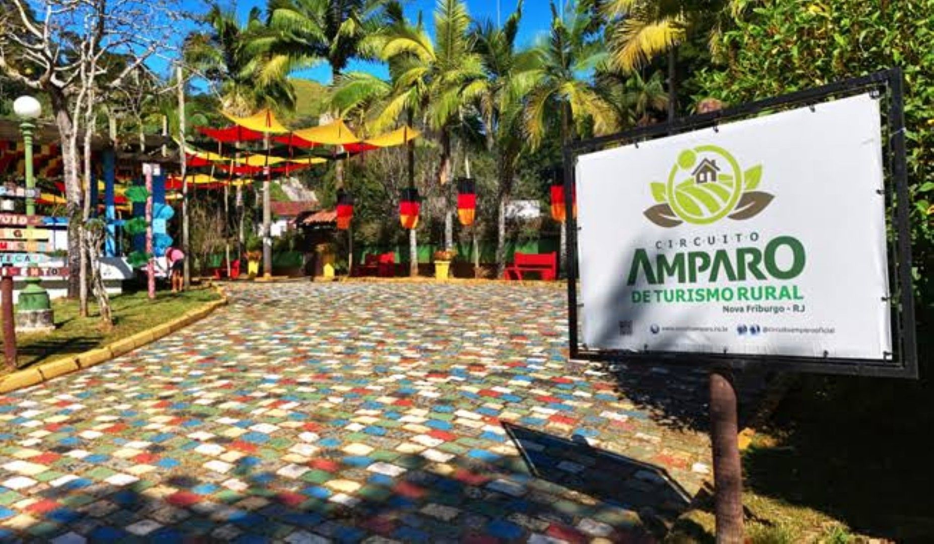 Placa indicativa do Amparo Ecoturismo Rural ao lado de uma praça de paralelepípedos colorida, com palmeiras e lanternas vermelhas.
