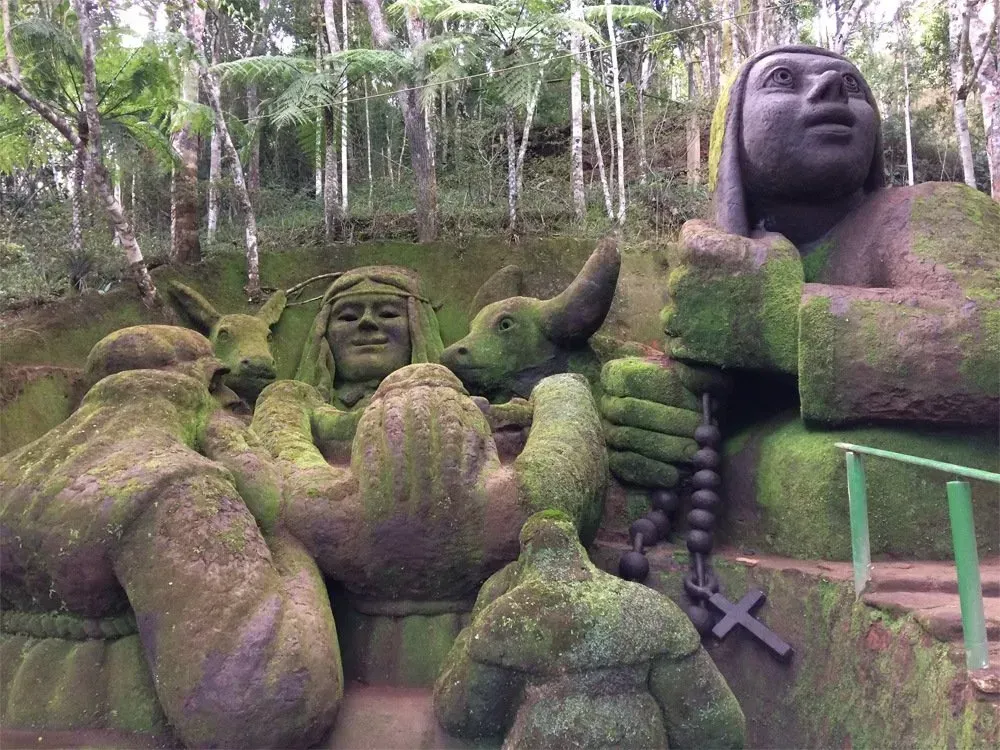 Estátuas de pedra cobertas de musgo em uma floresta, incluindo uma grande figura sentada e figuras menores com os braços erguidos.
