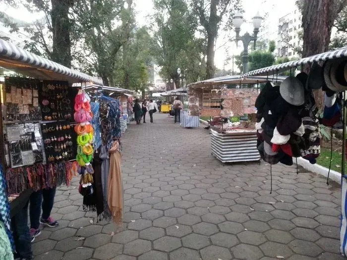 Mercado ao ar livre com calçada repleta de barracas de roupas e chapéus pendurados sob as árvores.