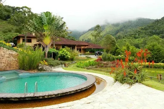 Resort tropical com piscina turquesa, flores vermelhas e montanhas verdejantes envoltas em névoa ao fundo.