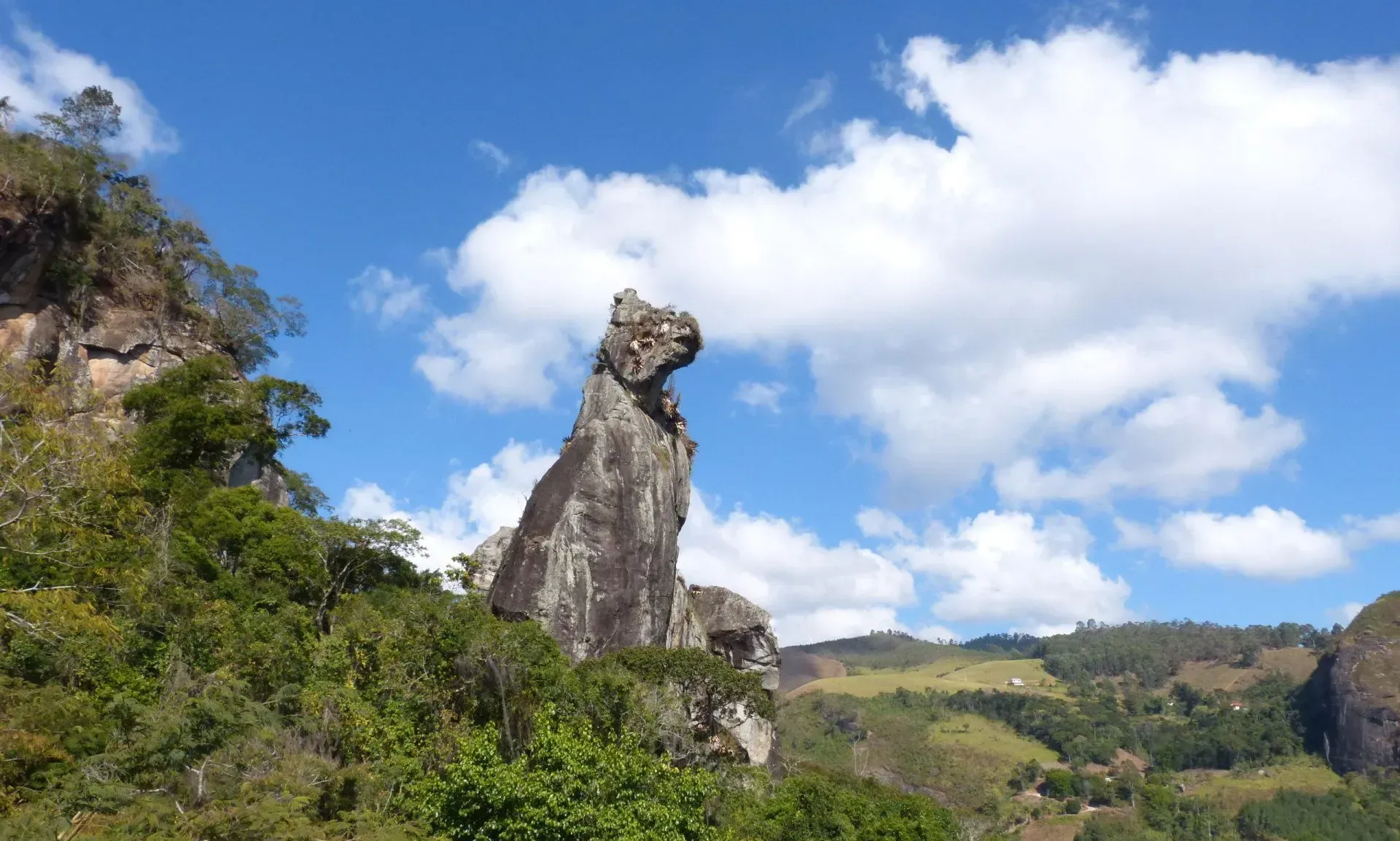 Alto monólito rochoso em meio a colinas verdejantes sob um céu azul brilhante com nuvens.
