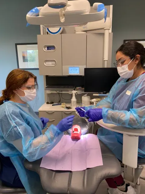 Dentist and assistant working on a patient’s teeth in a dental clinic, with a pink bib and mask-covered faces.
