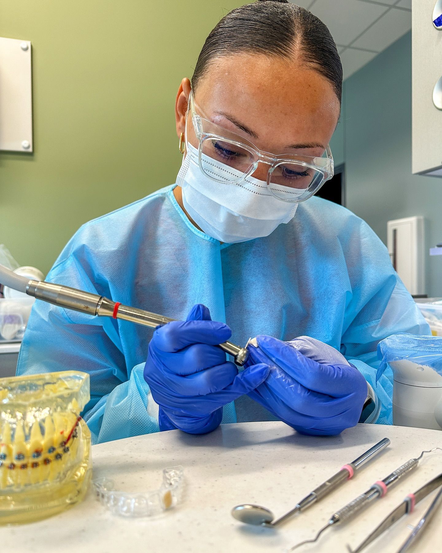 Dental hygienist in blue PPE cleaning a patient’s tooth with a dental tool and mirror nearby