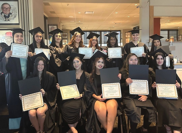 Group of graduates in caps and gowns holding diplomas and certificates at an indoor ceremony.