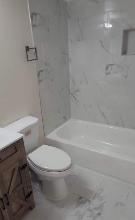 Modern bathroom with marble tile, white bathtub, toilet, and wood vanity.