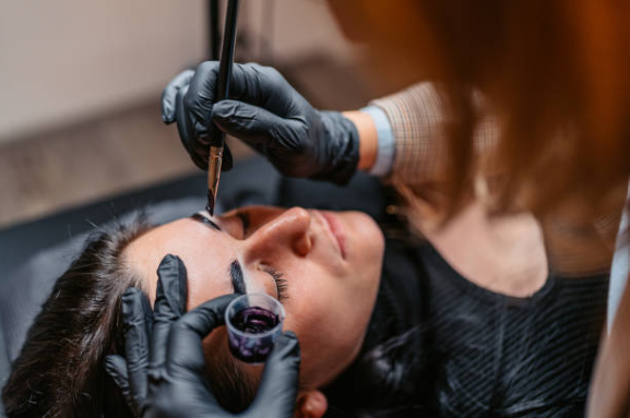 Eyelash technician applying mascara to a client lying down with eyes closed, using black gloves.