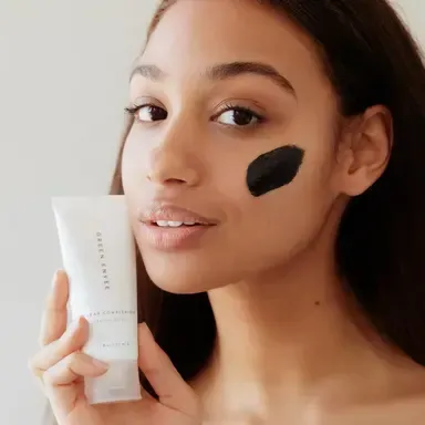 Woman applying black face mask while holding a white skincare tube against a light background